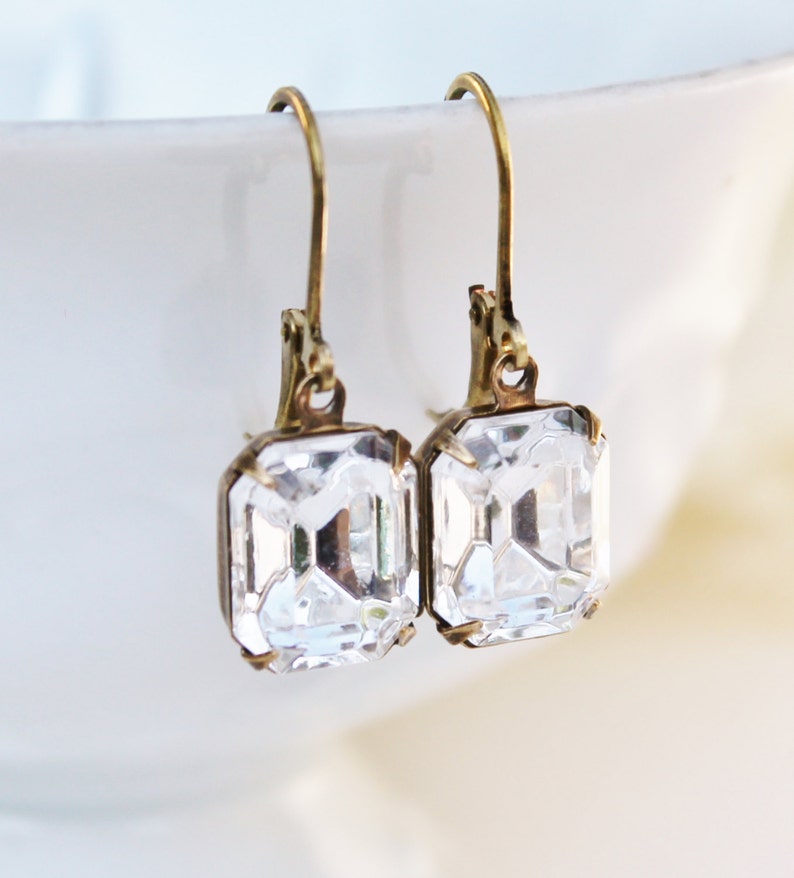 May include: A pair of vintage-style earrings with rectangular, clear crystal stones set in antique gold-tone metal. The earrings have a lever-back closure and are displayed against a soft, blurred background.