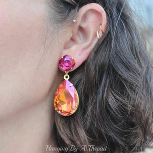 May include: A pair of dangle earrings featuring a teardrop-shaped orange and pink crystal, with a smaller pink crystal above it. The earrings are gold-toned.