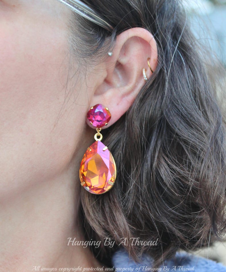 May include: A pair of dangle earrings featuring a teardrop-shaped orange and pink crystal, with a smaller pink crystal above it. The earrings are gold-toned.