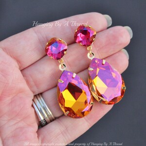 May include: A pair of dangle earrings with a teardrop-shaped crystal in a gradient of orange and pink. The earrings have a smaller round crystal in pink at the top.