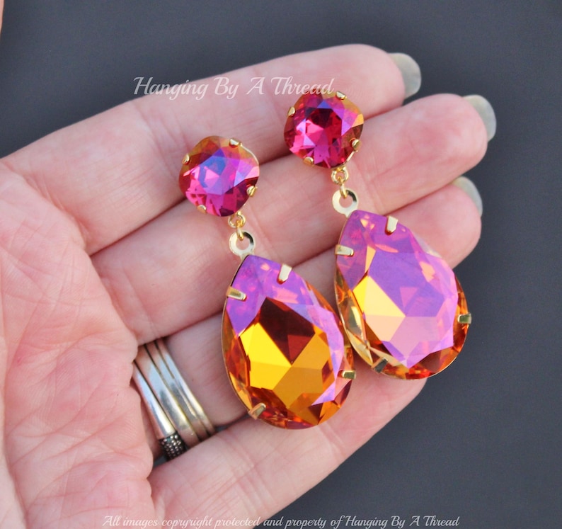 May include: A pair of dangle earrings with a teardrop-shaped crystal in a gradient of orange and pink. The earrings have a smaller round crystal in pink at the top.