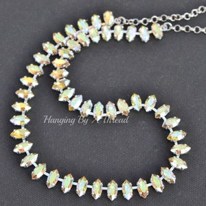 May include: A silver-toned necklace with iridescent, marquise-shaped crystals. The crystals are arranged in a continuous line, creating a sparkling, elegant design. The necklace is on a black background. Text on the image reads "Hanging By A Thread".