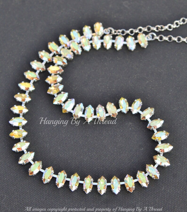 May include: A silver-toned necklace with iridescent, marquise-shaped crystals. The crystals are arranged in a continuous line, creating a sparkling, elegant design. The necklace is on a black background. Text on the image reads "Hanging By A Thread".