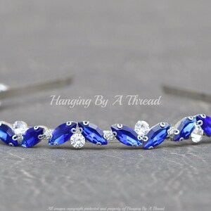 Pu&ograve; includere: Un cerchietto argentato con una fila di strass blu e trasparenti a forma di marquise. Il cerchietto &egrave; decorato con il testo "Hanging By A Thread".