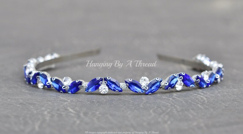 Pu&ograve; includere: Un cerchietto argentato con una fila di strass blu e trasparenti a forma di marquise. Il cerchietto &egrave; decorato con il testo "Hanging By A Thread".