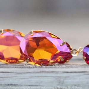 May include: A pair of dangle earrings featuring a teardrop-shaped orange and pink crystal at the top and a smaller round red crystal at the bottom. The earrings are made with gold-toned metal.