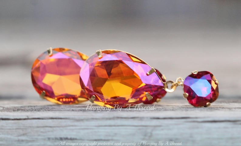 May include: A pair of dangle earrings featuring a teardrop-shaped orange and pink crystal at the top and a smaller round red crystal at the bottom. The earrings are made with gold-toned metal.
