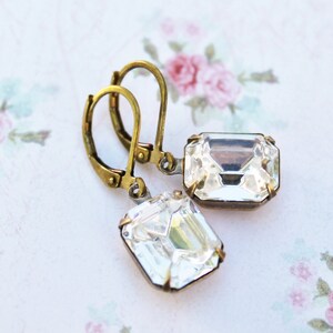 May include: A pair of vintage-style earrings with clear, rectangular crystals set in antique brass-colored metal. The earrings have lever-back closures and are displayed on a floral patterned background.