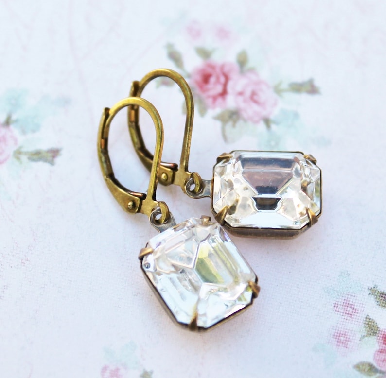 May include: A pair of vintage-style earrings with clear, rectangular crystals set in antique brass-colored metal. The earrings have lever-back closures and are displayed on a floral patterned background.