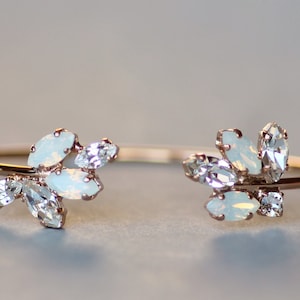 May include: A delicate open cuff bracelet with a rose gold finish. The bracelet features clusters of opalescent and clear crystals, creating a floral design. The crystals are set in a prong setting.