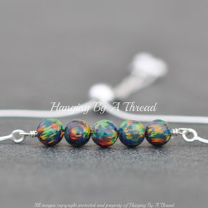May include: A silver adjustable bracelet with a row of five iridescent black beads with green and orange accents. The bracelet is made by Hanging By A Thread.