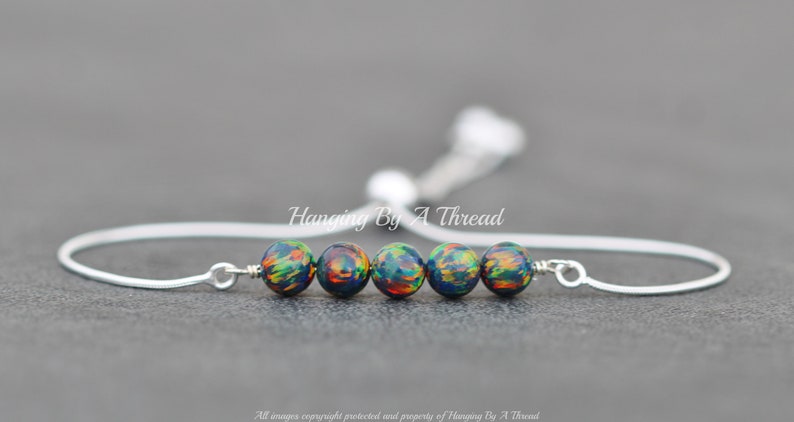 May include: A silver adjustable bracelet with a row of five iridescent black beads with green and orange accents. The bracelet is made by Hanging By A Thread.