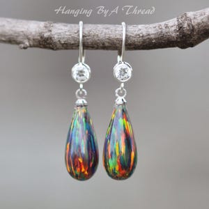May include: A pair of drop earrings featuring teardrop-shaped black opals with vibrant flashes of red, green, and yellow. Each earring has a clear round stone set in silver above the opal, suspended from a silver hook. The text Hanging By A Thread is visible.