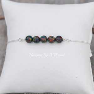 May include: A silver chain bracelet with six dark green and orange opal beads. The bracelet is on a white pillow with the text "Hanging By A Thread" printed on it.
