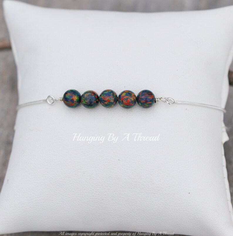 May include: A silver chain bracelet with six dark green and orange opal beads. The bracelet is on a white pillow with the text "Hanging By A Thread" printed on it.