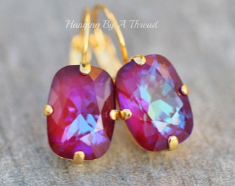 Puede incluir: Un par de pendientes de aro dorados con una piedra rectangular de color púrpura iridiscente. Los pendientes cuelgan de un gancho dorado.
