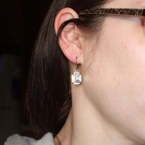 May include: A close-up of a clear, square-cut crystal earring with a gold-colored metal setting. The earring is hanging from a gold-colored hook. The earring is worn on the ear of a person.