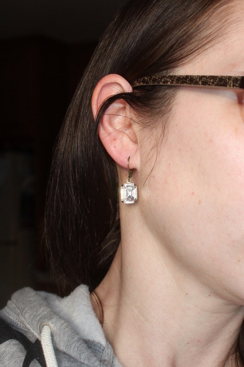 May include: A close-up of a clear, square-cut crystal earring with a gold-colored metal setting. The earring is hanging from a gold-colored hook. The earring is worn on the ear of a person.