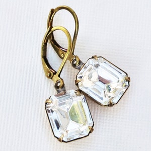 May include: A pair of leverback earrings featuring large, clear, emerald-cut crystals. The crystals are set in a gold-toned metal frame. The earrings have a vintage or antique aesthetic, with a simple, elegant design.