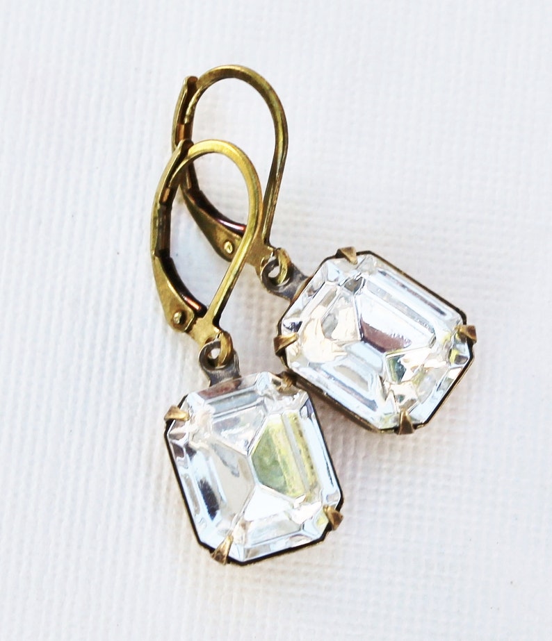 May include: A pair of leverback earrings featuring large, clear, emerald-cut crystals. The crystals are set in a gold-toned metal frame. The earrings have a vintage or antique aesthetic, with a simple, elegant design.