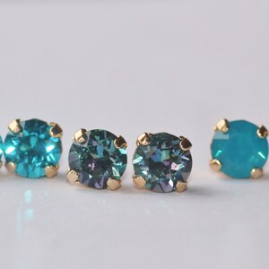 May include: A set of six stud earrings with gold settings and round, faceted gemstones. Two earrings feature a teal blue colour, while the other two feature a dark teal blue with a hint of purple.