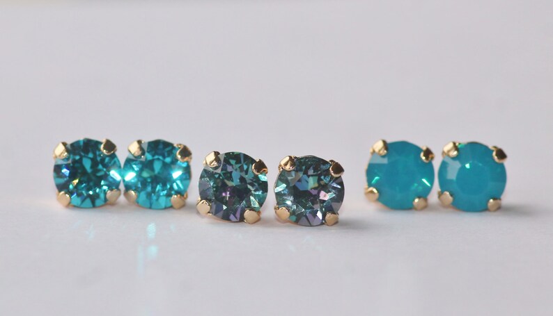 May include: A set of six stud earrings with gold settings and round, faceted gemstones. Two earrings feature a teal blue colour, while the other two feature a dark teal blue with a hint of purple.