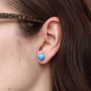 May include: A close-up of a person's ear with a blue opal stud earring. The earring is a round, flat shape with a light blue color and a shimmery finish.