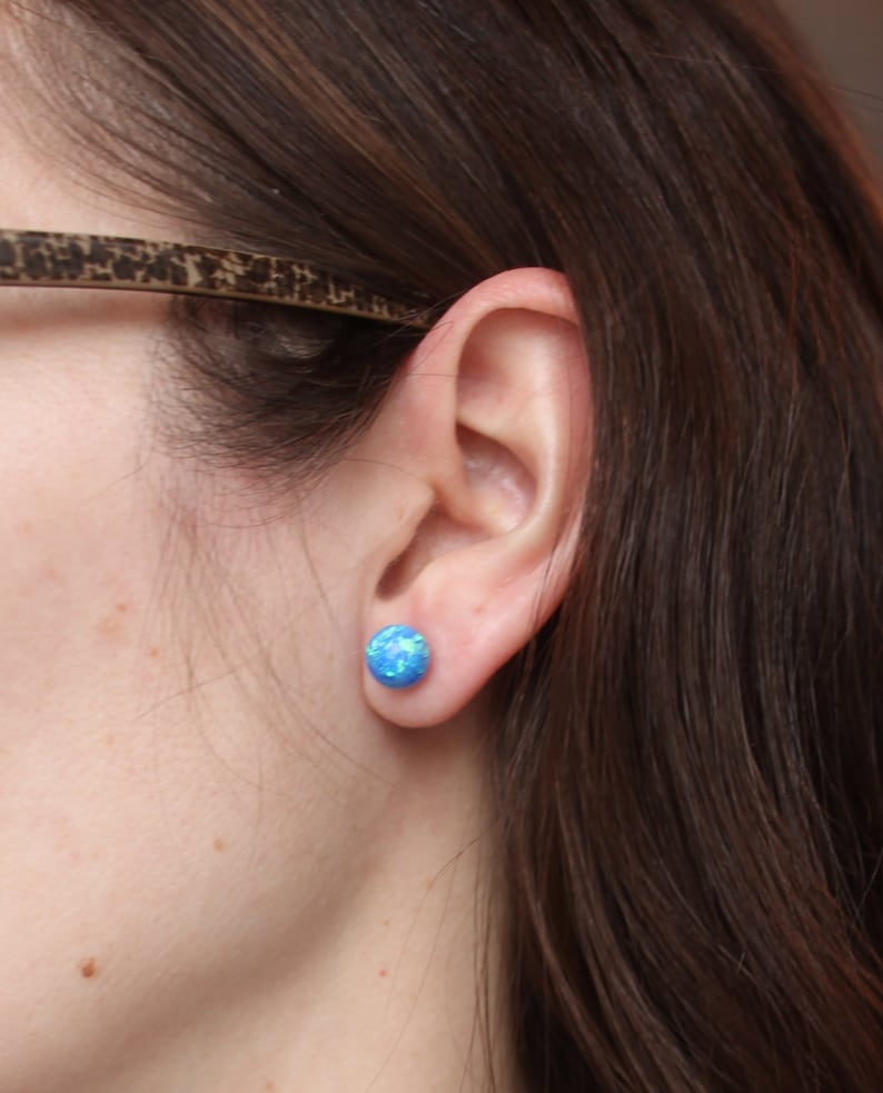 May include: A close-up of a person's ear with a blue opal stud earring. The earring is a round, flat shape with a light blue color and a shimmery finish.