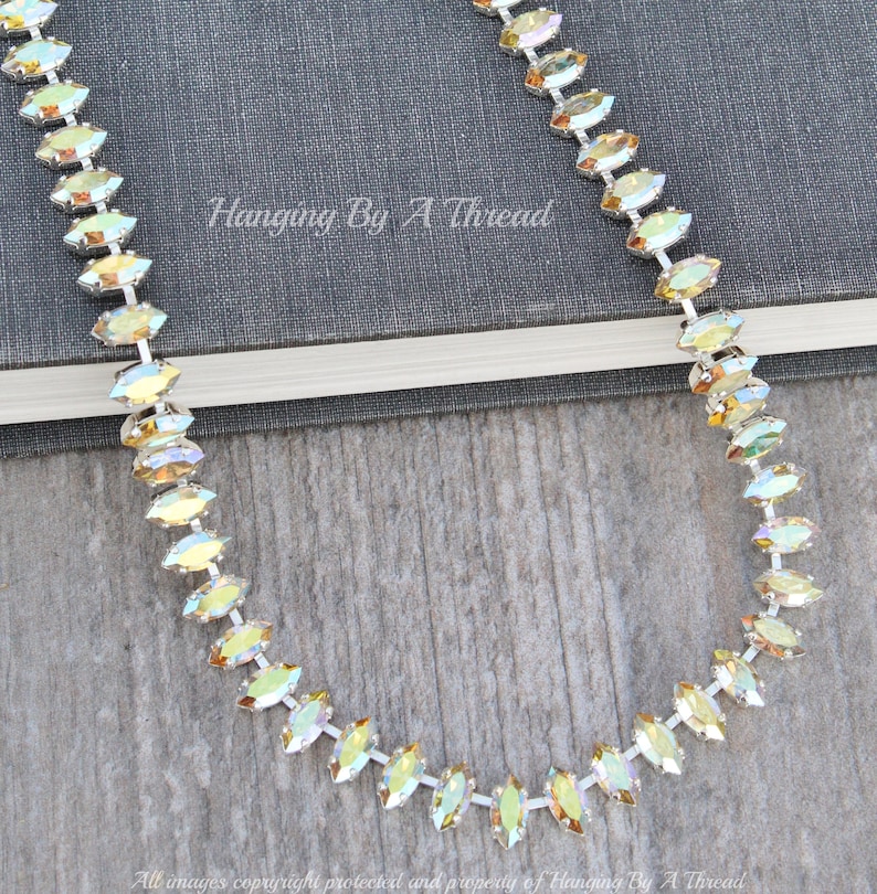 May include: A silver necklace with iridescent, marquise-shaped crystals. The necklace is displayed on a wooden surface with a book in the background. The text "Hanging By A Thread" is visible.