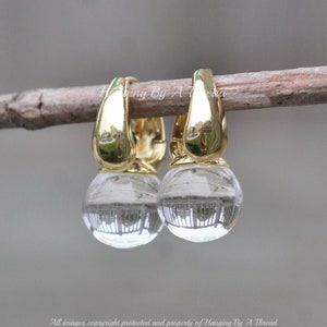 May include: A pair of gold-colored hoop earrings with clear, spherical crystal drops. The earrings are hanging on a brown twig. The text "Hanging By A Thread" is visible in the image.