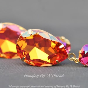 May include: A pair of dangle earrings featuring teardrop-shaped orange and pink crystals. The larger crystal is teardrop-shaped with a gold setting. The smaller crystal is square-shaped with a gold setting. The crystals are iridescent and sparkle.