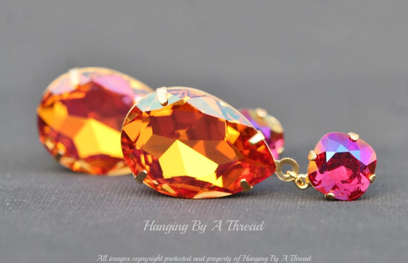 May include: A pair of dangle earrings featuring teardrop-shaped orange and pink crystals. The larger crystal is teardrop-shaped with a gold setting. The smaller crystal is square-shaped with a gold setting. The crystals are iridescent and sparkle.