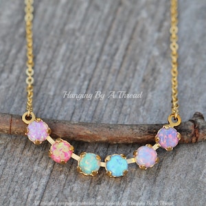 May include: A gold chain necklace with a rainbow of pastel opal stones. The stones are set in gold and are arranged in a curved line. The necklace is hanging on a wooden surface.