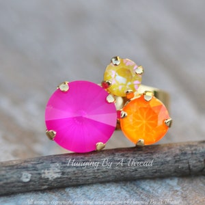 May include: A gold-toned ring featuring three colorful, faceted gemstones. The ring has a large, hot pink stone, a smaller orange stone, and a yellow stone. The ring is set on a wooden surface. The text "Hanging By A Thread" is visible.