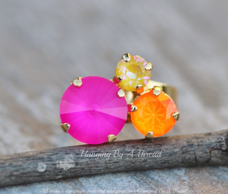 May include: A gold-toned ring featuring three colorful, faceted gemstones. The ring has a large, hot pink stone, a smaller orange stone, and a yellow stone. The ring is set on a wooden surface. The text "Hanging By A Thread" is visible.