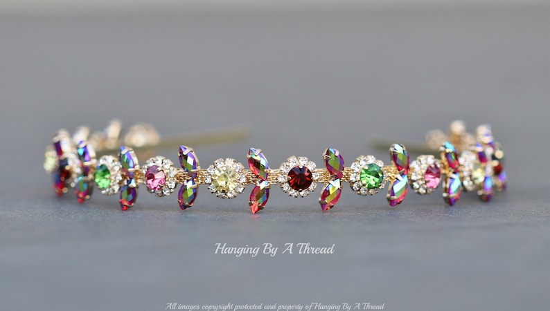 May include: A gold headband with a row of colorful iridescent rhinestones. The rhinestones are set in a floral design with small clear rhinestones surrounding each larger colored rhinestone.