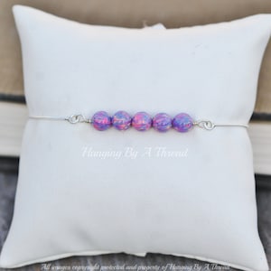 Puede incluir: Una pulsera de plata con una fila de seis cuentas de &oacute;palo p&uacute;rpura y rosa. La pulsera est&aacute; sobre una almohada blanca.