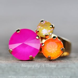 May include: A gold-toned ring featuring three large, faceted gemstones in vibrant colors: hot pink, orange, and yellow. The ring has a simple band and the gemstones are set with small gold prongs. The image includes the text "Hanging By A Thread".
