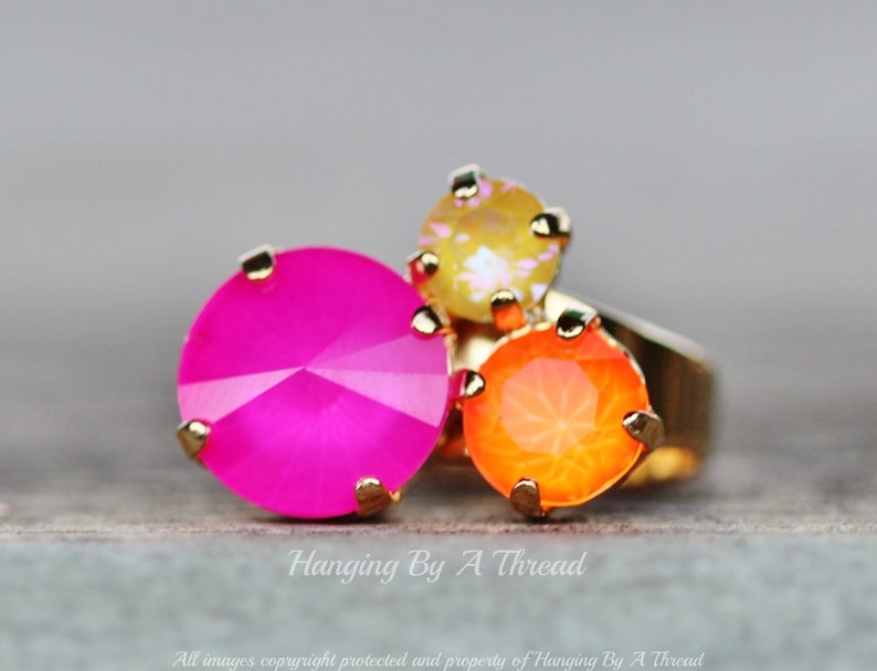 May include: A gold-toned ring featuring three large, faceted gemstones in vibrant colors: hot pink, orange, and yellow. The ring has a simple band and the gemstones are set with small gold prongs. The image includes the text "Hanging By A Thread".