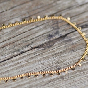 Puede incluir: Un collar de cadena de oro con peque&ntilde;os colgantes redondos. El collar est&aacute; sobre una superficie de madera.