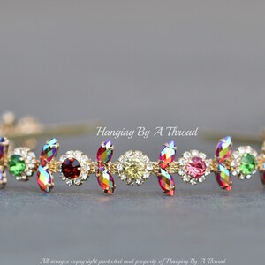 May include: A gold headband with a row of iridescent, multi-colored, teardrop-shaped crystals. The crystals are set in a gold metal setting and are surrounded by smaller clear crystals. The headband is called "Hanging By A Thread".