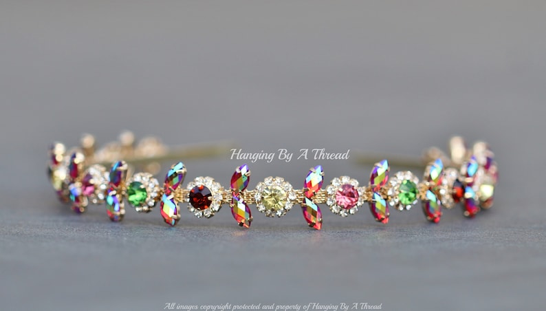 May include: A gold headband with a row of iridescent, multi-colored, teardrop-shaped crystals. The crystals are set in a gold metal setting and are surrounded by smaller clear crystals. The headband is called "Hanging By A Thread".