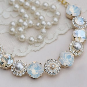 May include: A gold-toned necklace with large, iridescent, square and round crystals, and smaller, round, pearl-like beads. A separate strand of white pearls is also visible. The jewelry is set against a white lace background.