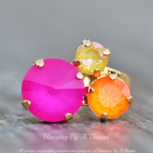 May include: A gold-toned ring featuring three colorful, faceted gemstones. The ring has a large, bright pink stone, an orange stone, and a yellow stone. The ring is set against a blurred background.