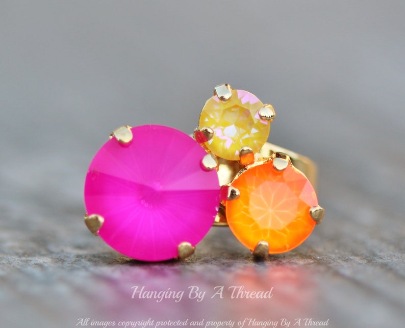 May include: A gold-toned ring featuring three colorful, faceted gemstones. The ring has a large, bright pink stone, an orange stone, and a yellow stone. The ring is set against a blurred background.