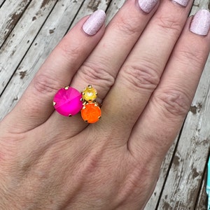 May include: A gold-toned ring featuring three large, colorful gemstones. The ring has a large, hot pink gemstone, a smaller yellow gemstone, and an orange gemstone. The ring is set on a hand with light pink glitter nail polish.