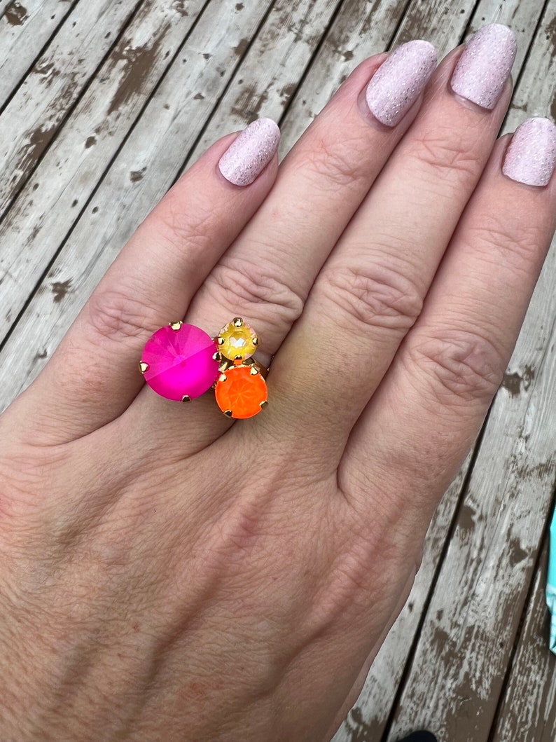May include: A gold-toned ring featuring three large, colorful gemstones. The ring has a large, hot pink gemstone, a smaller yellow gemstone, and an orange gemstone. The ring is set on a hand with light pink glitter nail polish.
