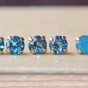 May include: Five pairs of small, round, gold-toned stud earrings with blue gemstones. The gemstones are in two shades of blue, a darker teal and a lighter turquoise.