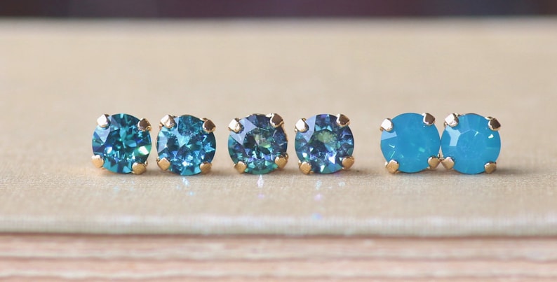 May include: Five pairs of small, round, gold-toned stud earrings with blue gemstones. The gemstones are in two shades of blue, a darker teal and a lighter turquoise.