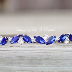 Pu&ograve; includere: Un braccialetto d'argento con strass a forma di marquise blu e trasparenti alternati. Il braccialetto &egrave; su una superficie di legno.